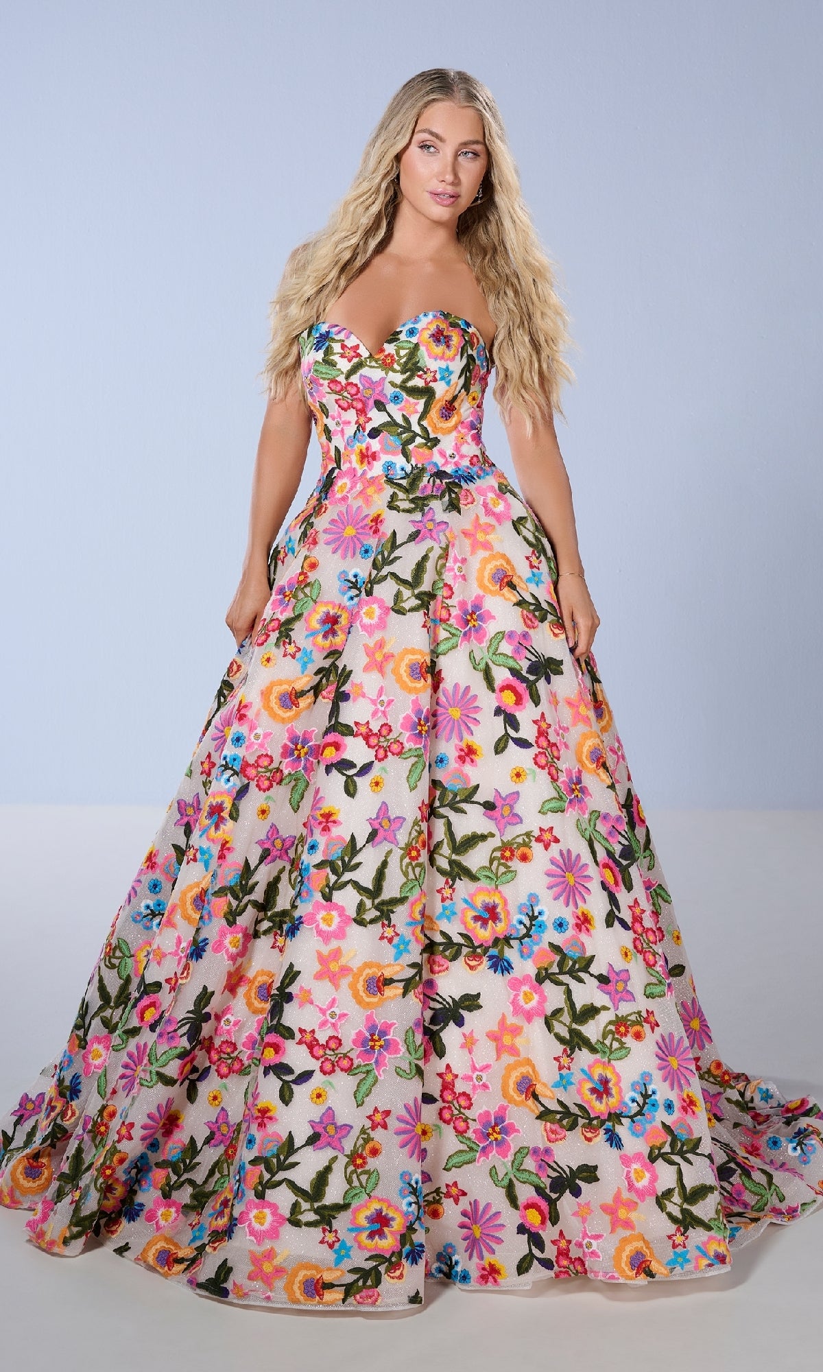 A woman models the Tiffany 16176 Floral Embroidered Prom Ball Gown, featuring a strapless sweetheart neckline and colorful floral embroidery, set against a light blue background.